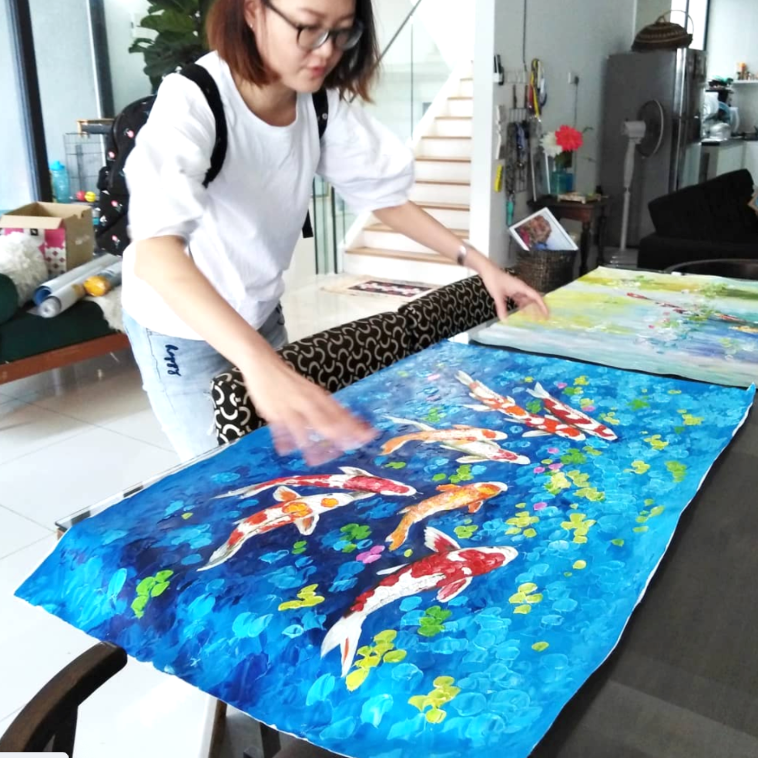 A woman with glasses and a white shirt is working on a colorful abstract painting spread out on a table, with a backpack on her shoulders in a bright, modern room.