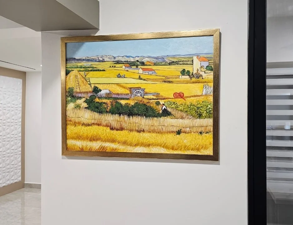 Landscape painting of farmland with fields, haystacks, buildings, mountains, and a tractor, framed on a white wall.