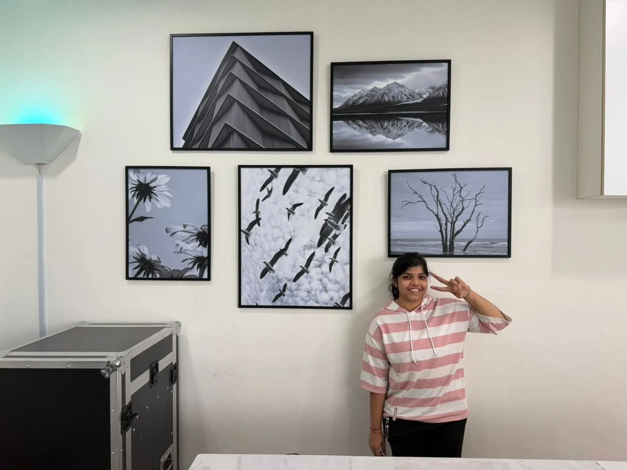 A young woman smiling and making a peace sign with her right hand, standing in front of a wall with six framed black-and-white landscape photographs, and standing near a floor lamp with a white shade in a room.