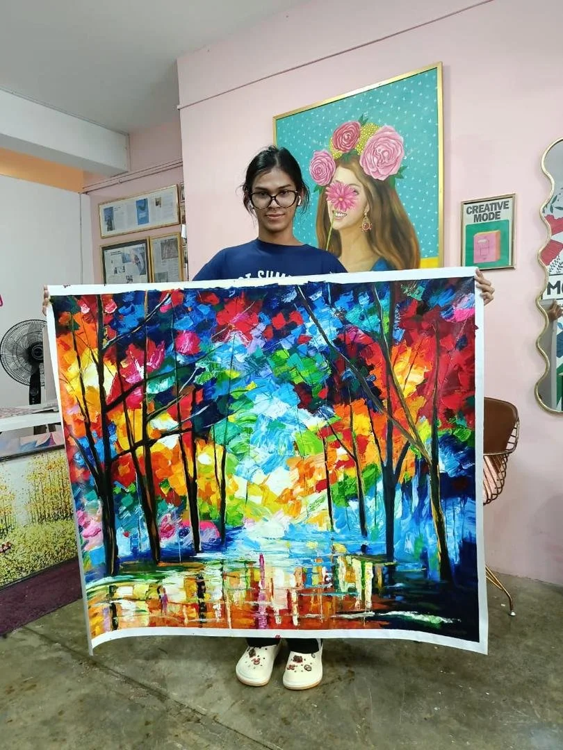 Young woman with glasses holding a colorful abstract landscape painting of a wooded scene with trees and a reflection in water.