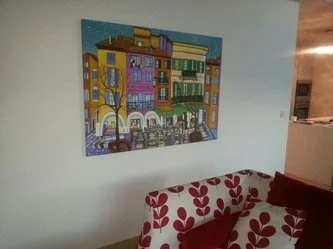 Colorful painting of a cityscape hanging on a light-colored wall above a sofa with red and white floral pattern cushions.