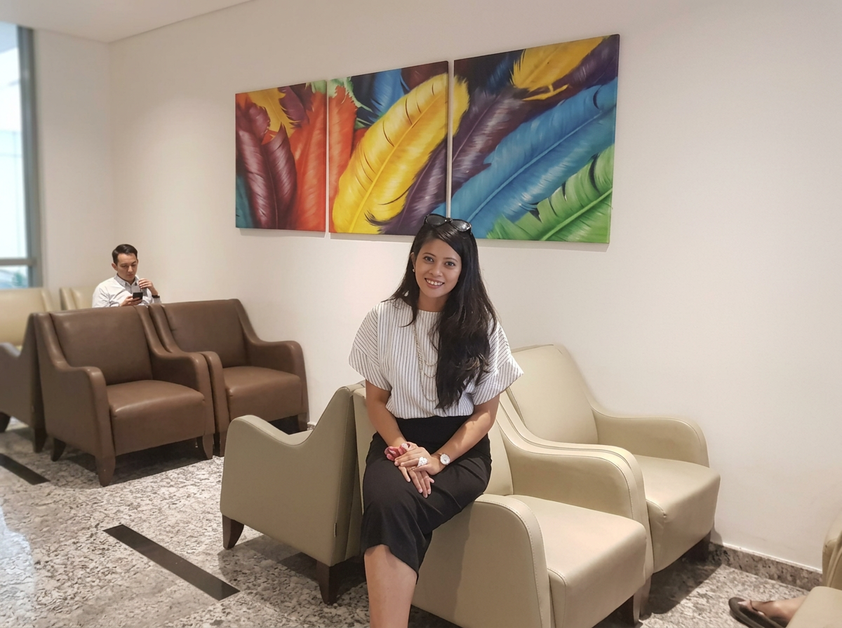 Smiling woman sitting on a beige armchair in a waiting area with colorful feather artwork on the wall behind her, and a man in the background looking at his phone.