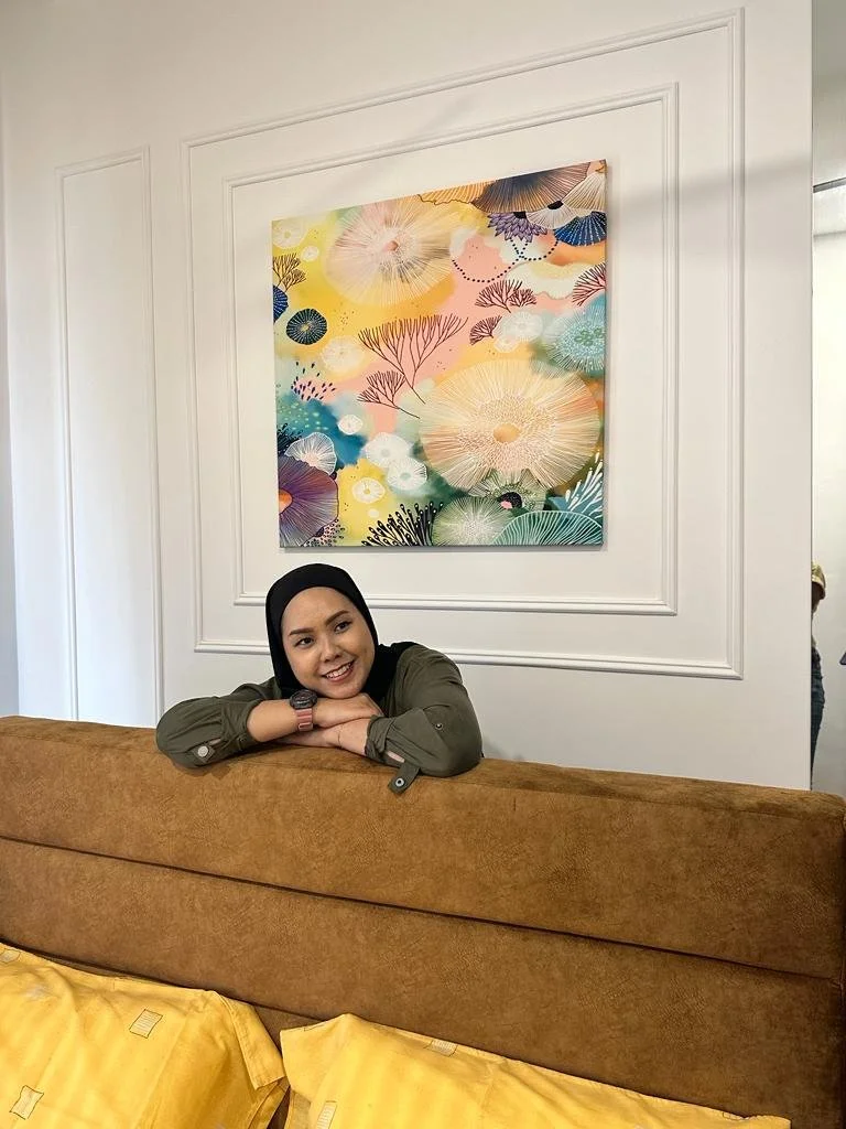 A woman smiling and resting her chin on her crossed arms above a brown sofa, with colorful abstract floral artwork on the white wall behind her.