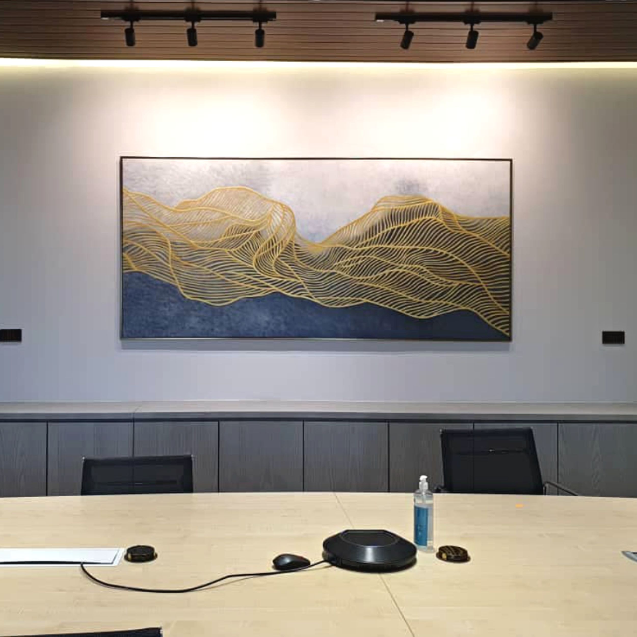 Conference room setup with a long table, chairs, a microphone, a water bottle, and electronic devices like a speakerphone and a remote control on the table. The background features a wall with a framed artwork and ceiling lights.