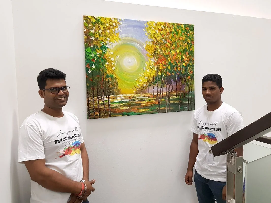 Two men standing beside an abstract landscape painting of a pathway through trees with autumn foliage and a bright sun in the sky, displayed on a white gallery wall.