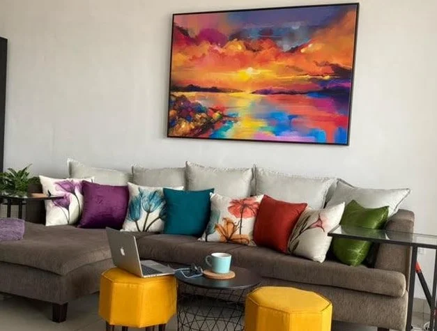 Living room with a gray sectional sofa with colorful throw pillows, a laptop, a mug, and two yellow ottomans in front of a large colorful landscape painting on the wall.