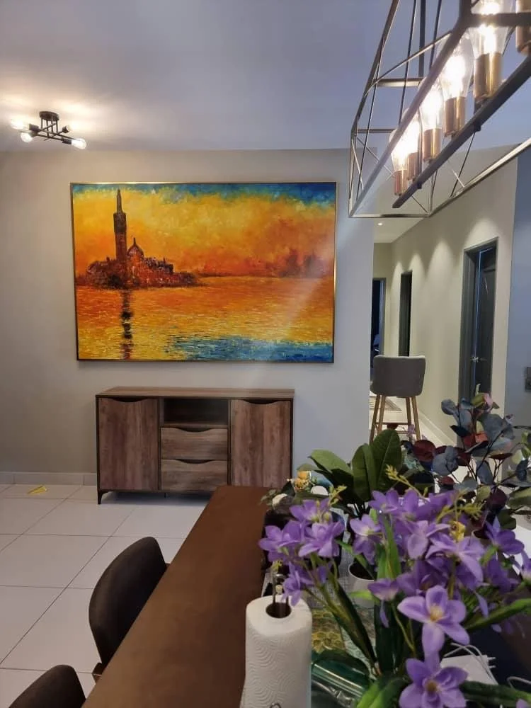 Living room wall with a large colorful painting of a sunset over water and a small wooden cabinet below it. A dining table with purple flowers and a paper towel roll is visible in the foreground. A modern light fixture hangs from the ceiling. There a