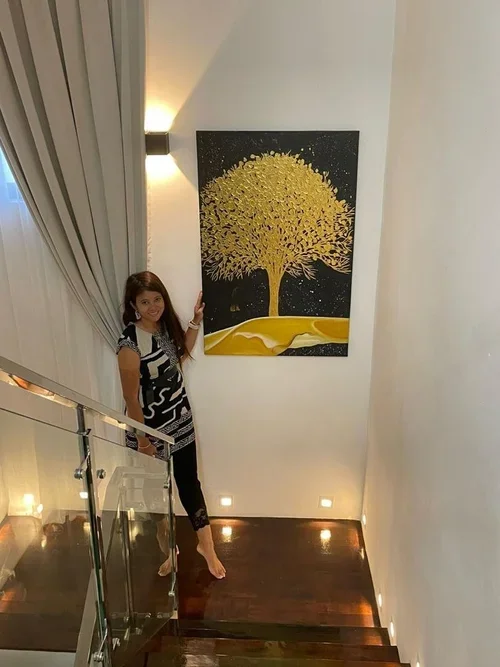 A woman with long hair wearing a black and white dress standing on a staircase inside a home or gallery, pointing at a large painting of a golden tree on a black background hanging on a white wall.