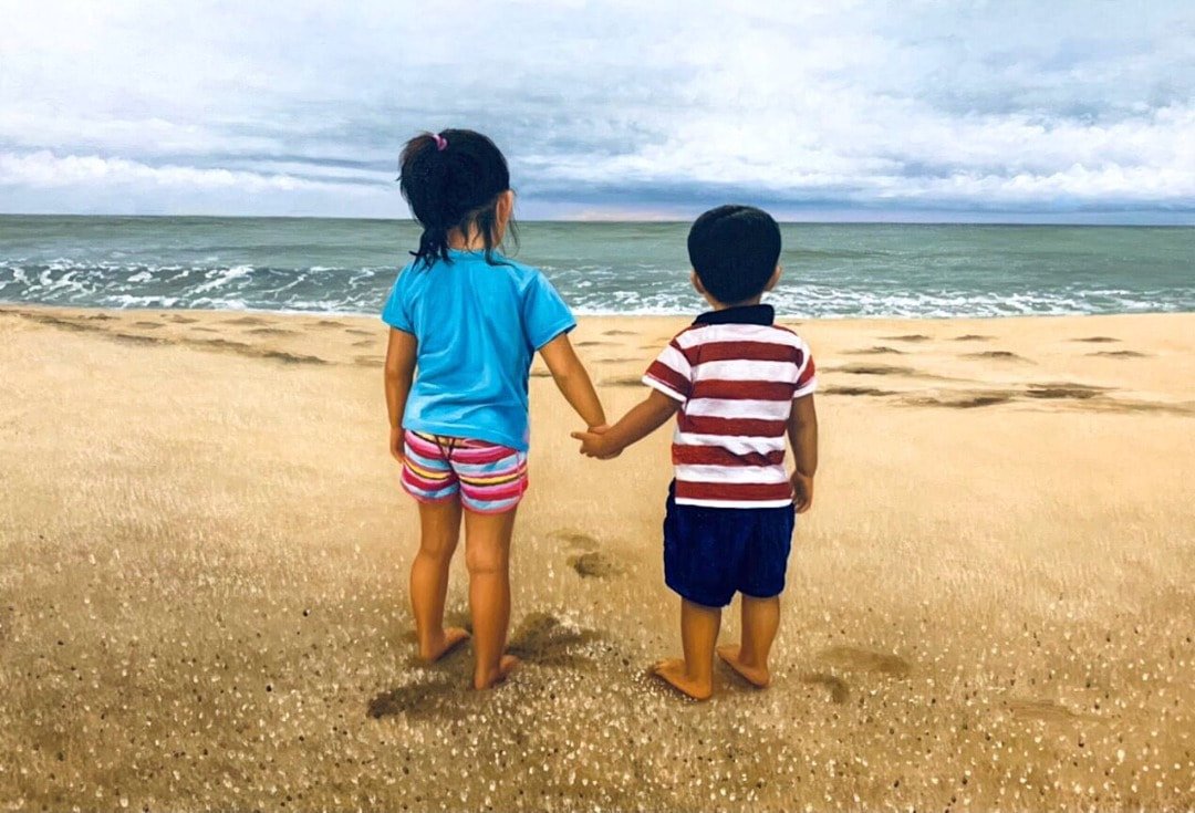 Two children, a girl and a boy, holding hands on a sandy beach facing the ocean, with cloudy sky above.