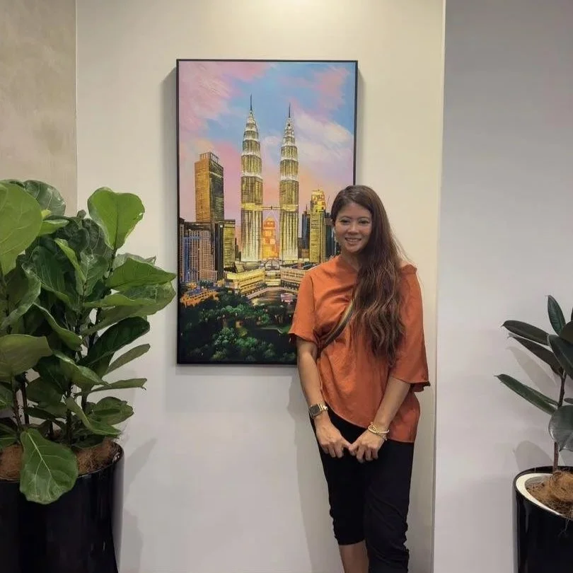 Woman with long brown hair wearing an orange shirt and black pants standing next to a painting of the Petronas Towers in Kuala Lumpur, Malaysia, with green potted plants on both sides.