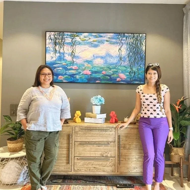 Two women standing on either side of a wooden sideboard in a living room, decorated with plants, colorful duck figurines, and a stack of books, with a large framed painting of water lilies hanging on the wall behind them.