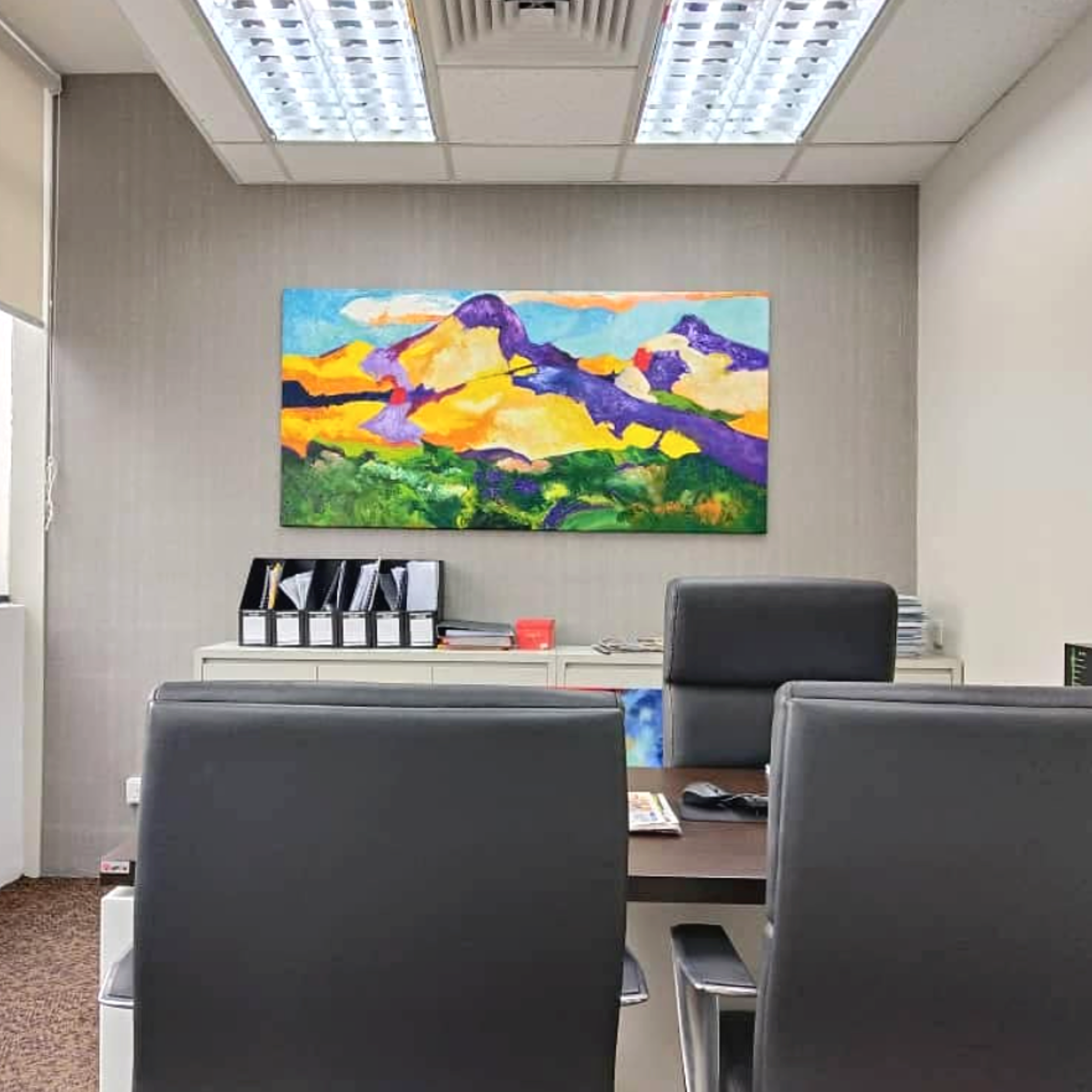 Office conference room with a colorful abstract painting on the wall, black binders on a white shelf, chairs, and a desk.