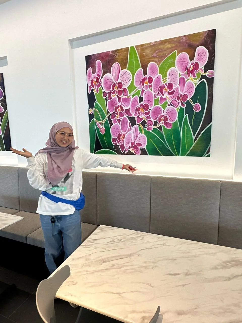A smiling woman wearing a light purple hijab and white shirt, standing indoors, gesturing towards a colorful painting of pink orchids with green leaves.