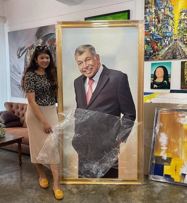 A woman standing next to a large framed portrait of a smiling man in a suit with gray hair, in an art gallery. Artwork hangs on the walls behind them.