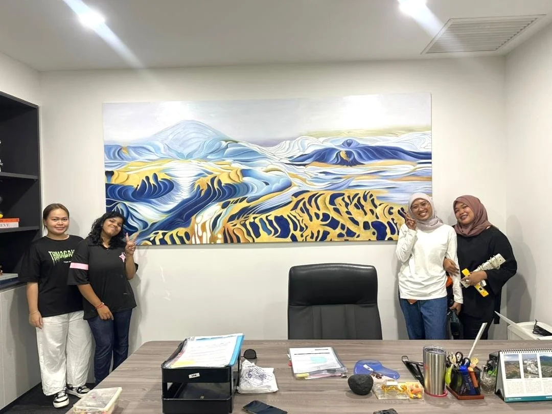 Four women are standing in an office room in front of a large abstract landscape painting featuring blue, yellow, and white colors. The women are smiling and posing, with two on each side of the room.