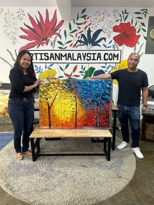 Two people standing next to a colorful landscape painting of trees during sunset or sunrise, with leaves changing colors, in an art studio with floral wall murals and a sign that reads 'ARTISANMALAYSIA.COM' behind them.