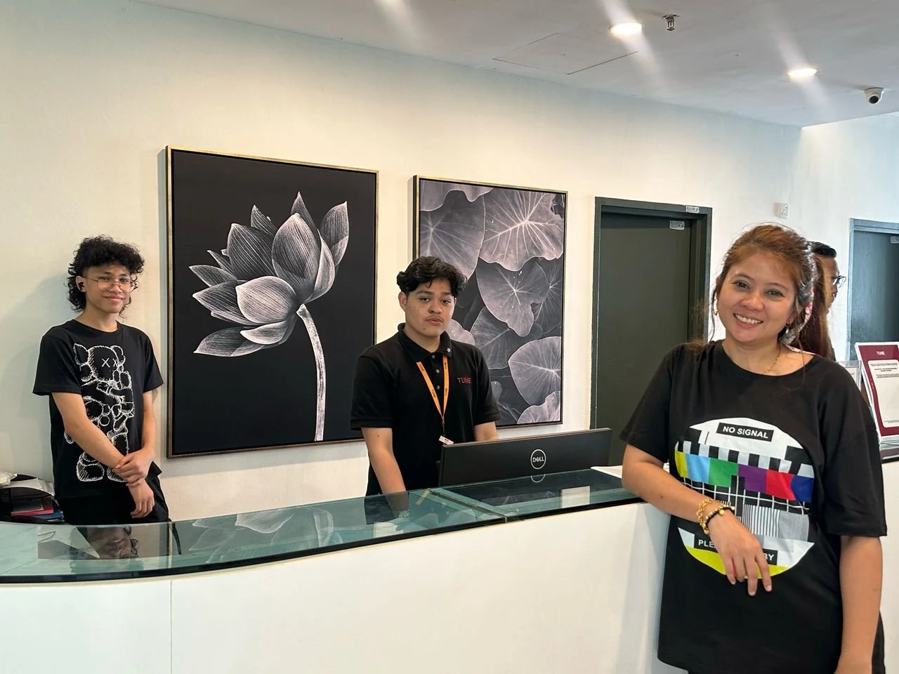 Three people standing behind a reception desk in an art gallery with black and white floral artwork on the wall. The woman in front is smiling, wearing a black T-shirt with a graphic design.