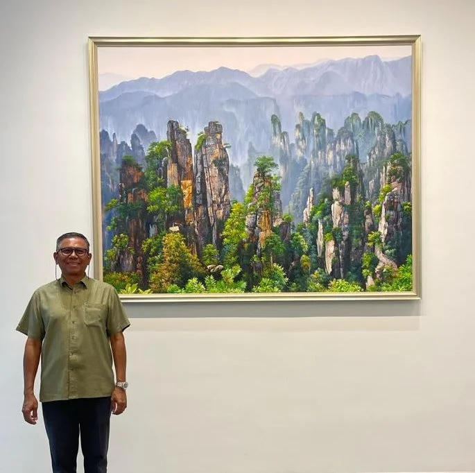 A man in a green shirt and glasses standing in front of a large framed landscape painting of jagged mountain peaks with trees and mist in the background.