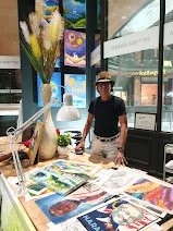 A person stands behind a table of various colorful books and magazines inside a store with large windows showing street view and bright lighting.