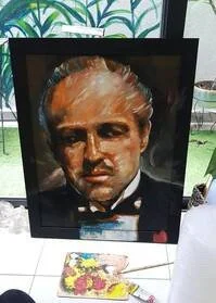 Framed portrait of a man with a serious expression, short hair, and wearing a dark suit. The frame is black, placed on a table with a colorful cartoon drawing nearby, with a window and plants in the background.