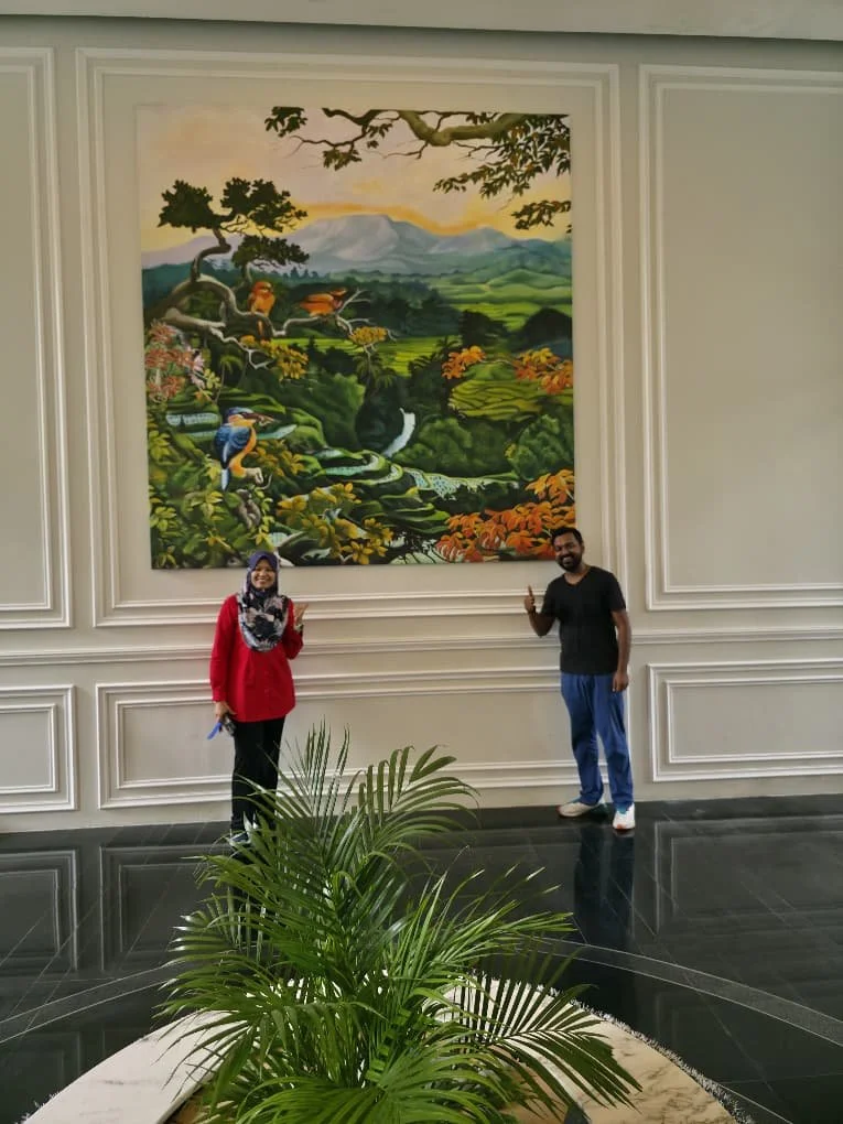 Two people standing in front of a large, colorful landscape painting of a natural scene with mountains, trees, waterfalls, and animals, inside a room with white walls and a black tiled floor.