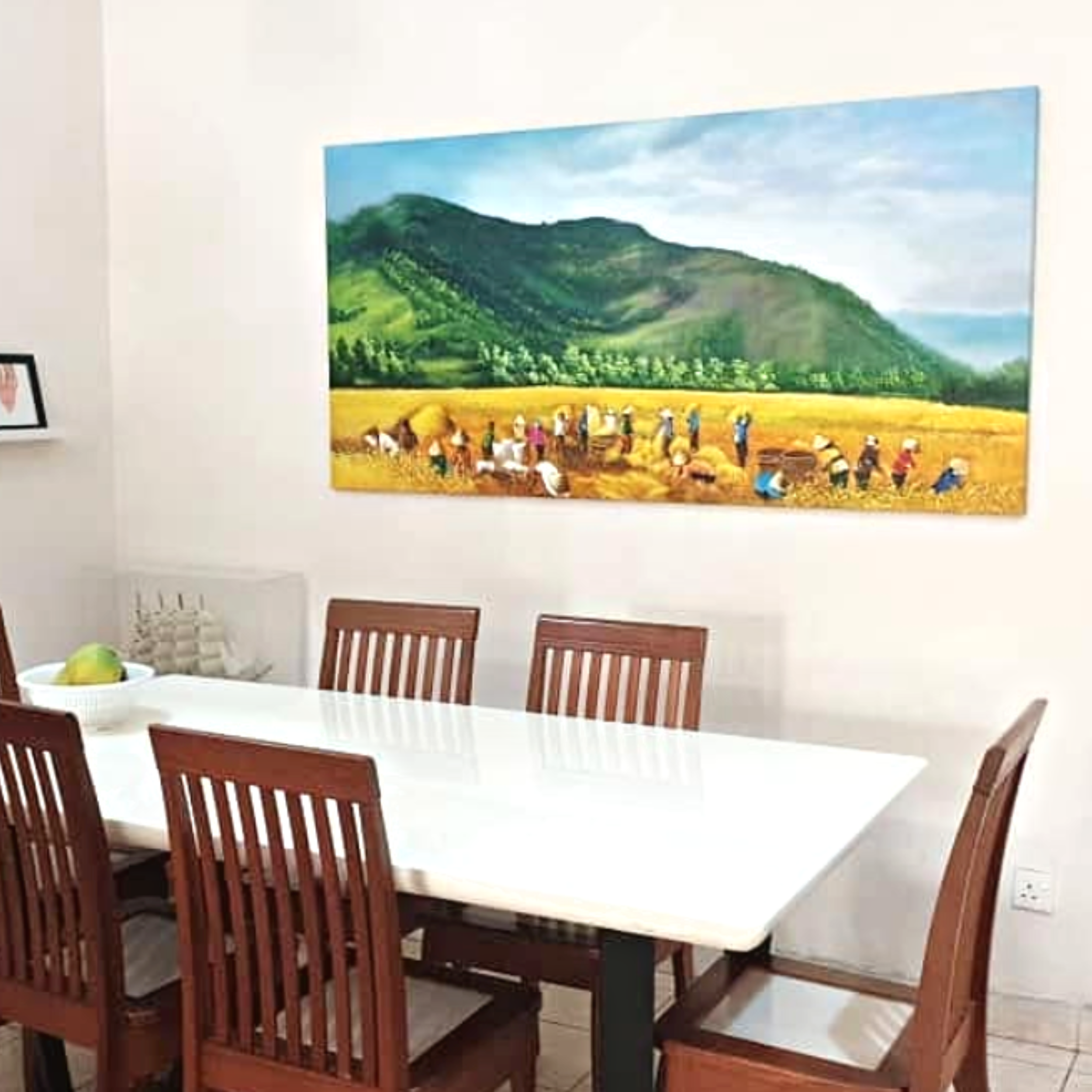 A large landscape painting of green hills, trees, and a partly cloudy sky hangs on the wall in a dining room. The room has a white wall and a white dining table with wooden chairs.
