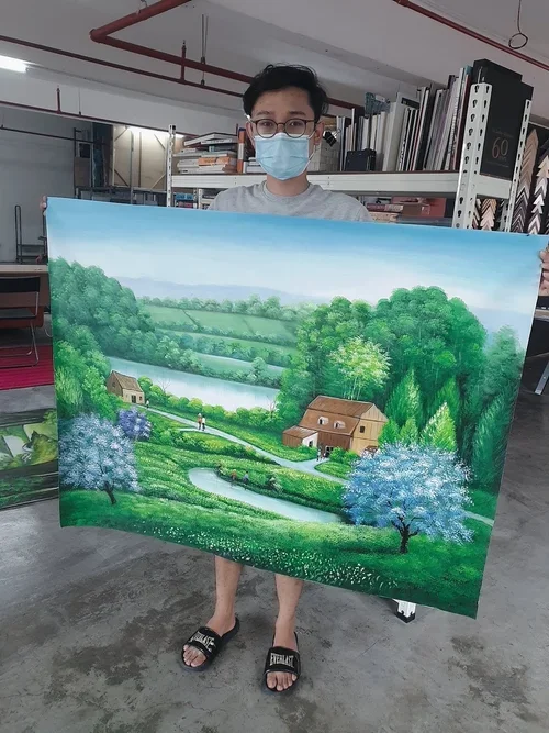 A person wearing glasses and a blue face mask holds a large landscape painting with green trees, a river, and houses, in an indoor space with shelves and books in the background.