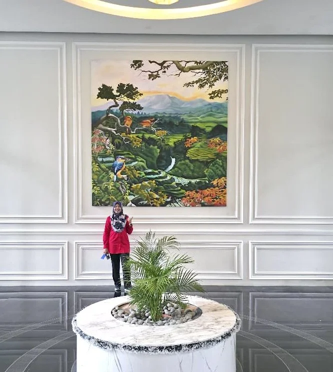 A woman in a red coat and scarf standing in front of a large landscape painting in a room with white paneled walls and a round marble table with a potted plant.