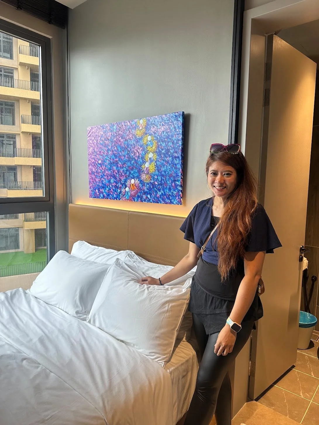 Woman with long brown hair standing next to a bed in a hotel room, smiling at the camera, with a colorful abstract painting on the wall behind her and a window showing a building outside.
