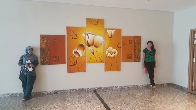 Two women standing on opposite sides of a multi-panel floral artwork on a white wall. One woman is wearing a hijab and holding papers, the other is in a striped shirt holding a pink bag.