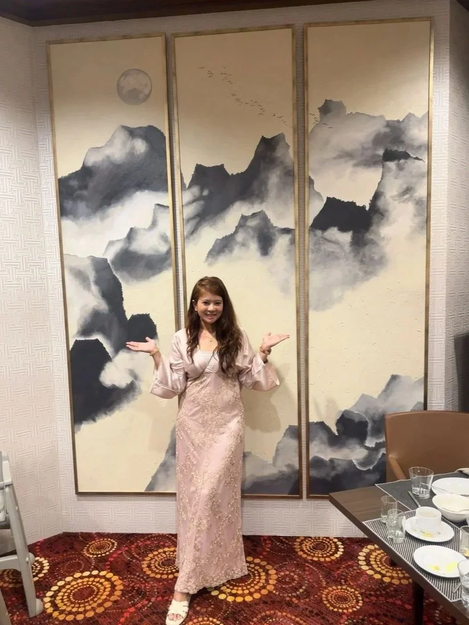 A woman in a pink lace dress standing in front of a large three-panel painting of mountains, with a smiling expression and her arms raised. The room has a patterned carpet and a table with dishes and glasses on the right side.