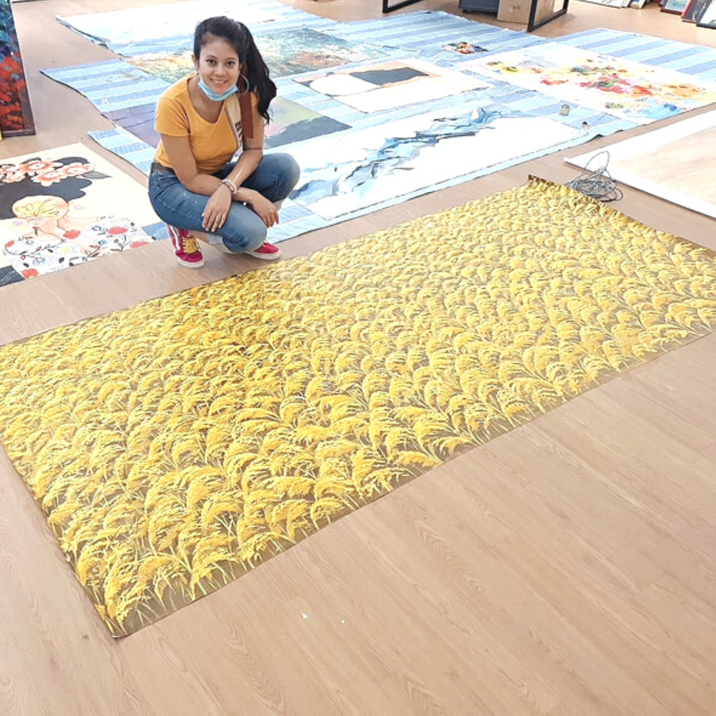 A young woman with dark hair, wearing a yellow shirt, jeans, and red shoes, sitting on a wooden floor surrounded by large colorful paintings or fabric artworks laid out flat.