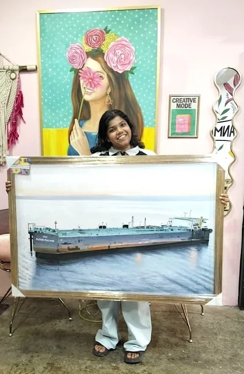 A young girl standing inside a colorful room, holding a framed picture of a large ship on the water. Behind her, there is a portrait of a woman with flowers over her face, and a framed sign that says "CREATIVE MODE". The wall is decorated with art an