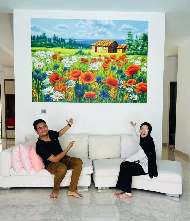 Two people sitting on a white sofa in a living room, smiling and pointing at a large colorful landscape painting on the wall. The painting depicts a field of flowers with a cottage, trees, and a blue sky.