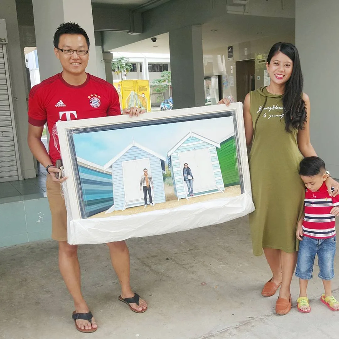 A family of three holding a framed painting outside a building. The man wears glasses and a red sports jersey, the woman wears a green dress, and the child is dressed in a red and white striped shirt with denim shorts.