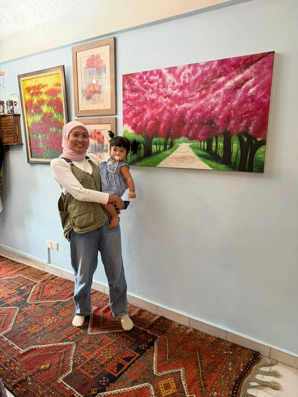 A woman wearing a pink hijab and glasses holding a young girl with a pacifier in her mouth, standing inside an art gallery. The woman is smiling and the girl is looking at the camera. Behind them are framed paintings of pink flowers and a pathway lin