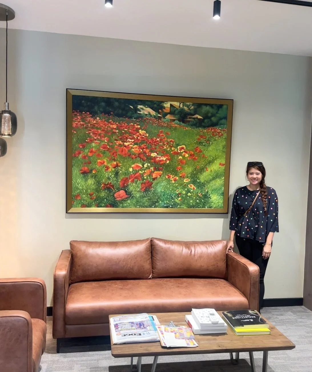 A woman standing next to a tan leather sofa in a living room with a large, framed painting of a lush green field filled with red, orange, and yellow flowers hanging on the wall behind her.