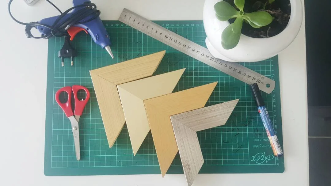Four small wooden picture frames on a green cutting mat, with scissors, hot glue gun, ruler, marker, and a potted plant nearby.