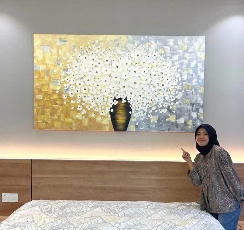 A woman wearing a patterned blouse and hijab smiling and pointing at a large painting of white flowers in a black vase, hanging above a bed in a modern bedroom.