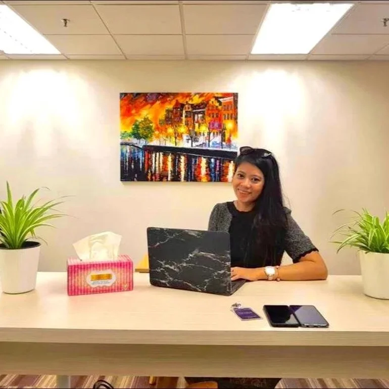 A woman sitting at a desk with a black marble-patterned laptop, tissue box, and two smartphones. There are two potted green plants on either side of the desk and a colorful cityscape painting on the wall behind her.