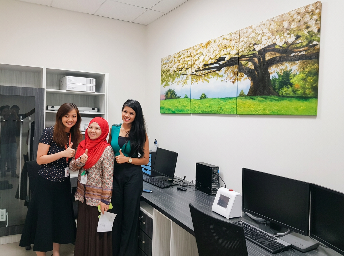 Three women standing in an office, smiling and giving thumbs up, with computer monitors and a large landscape painting of a tree on the wall.