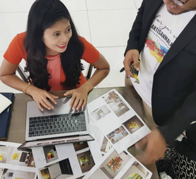 A young girl with long dark hair in a red shirt is smiling and pointing at a laptop and printed photos on a table, while an adult person standing next to her holds a phone and looks at the photos.