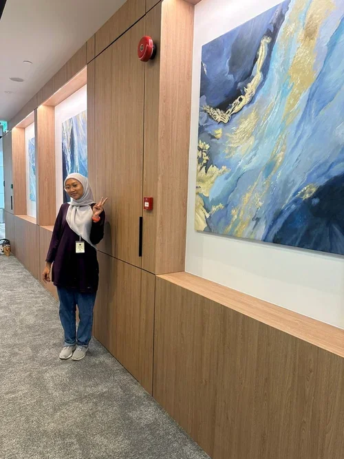A woman wearing a hijab and business attire standing in a hallway with wooden panels and blue abstract paintings, making a peace sign with her hand.