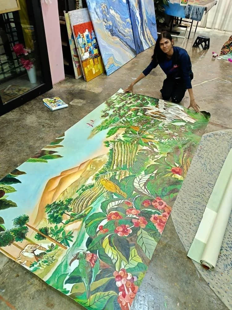 An artist kneeling on the floor, working on a large colorful painting of a lush landscape with trees, waterfalls, mountains, and animals, with other paintings visible in the background.