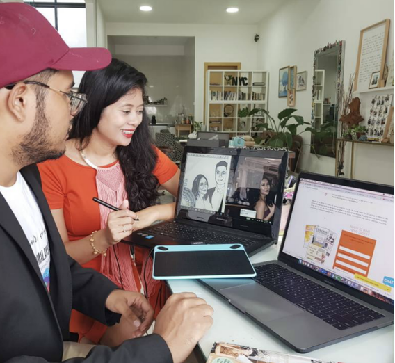 Two people sitting at a table with multiple laptops, engaged in a virtual meeting or presentation, in a brightly lit, modern room with bookshelves and artwork.