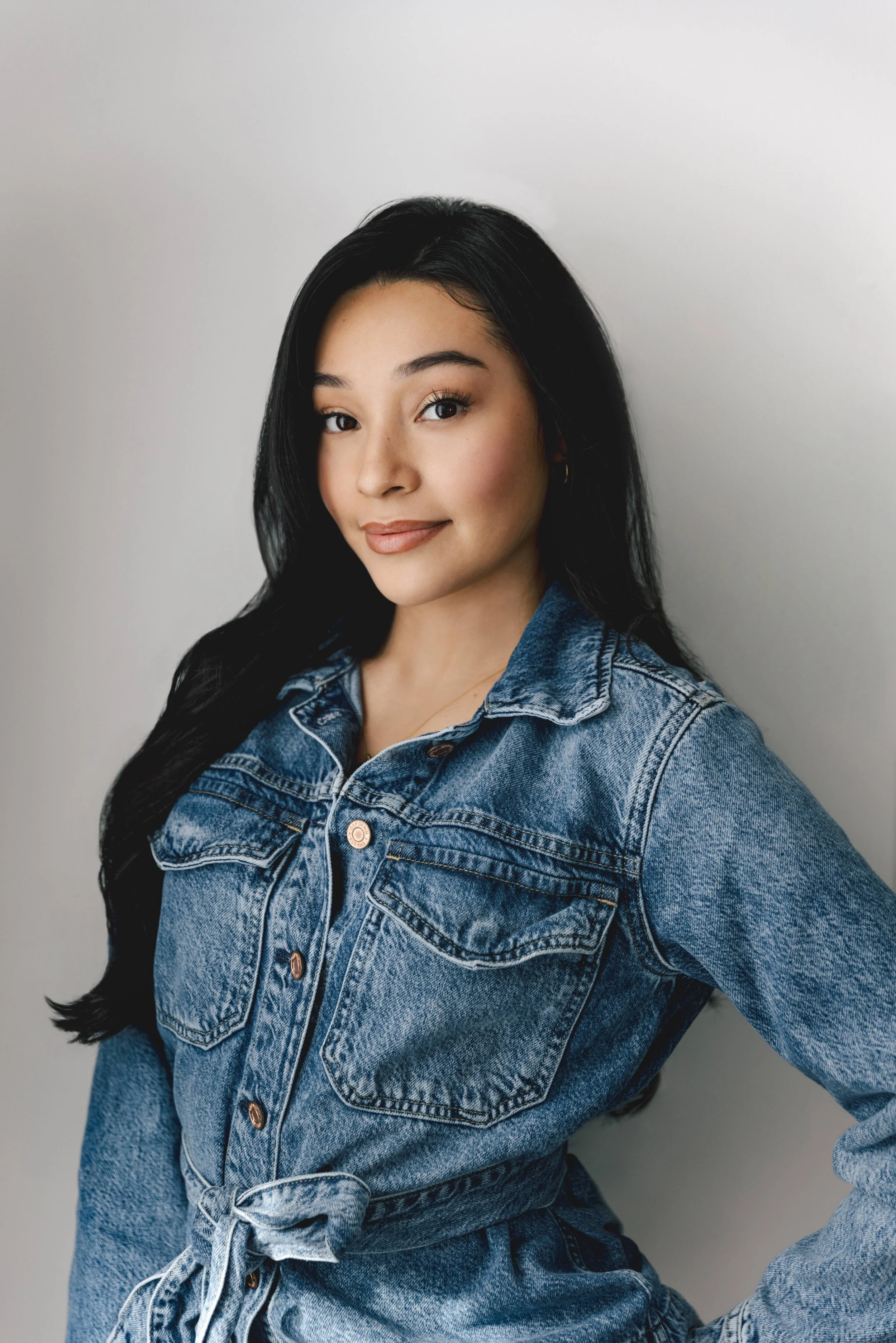 Portrait of a woman with long black hair wearing a denim jacket, standing against a plain, light-colored background.
