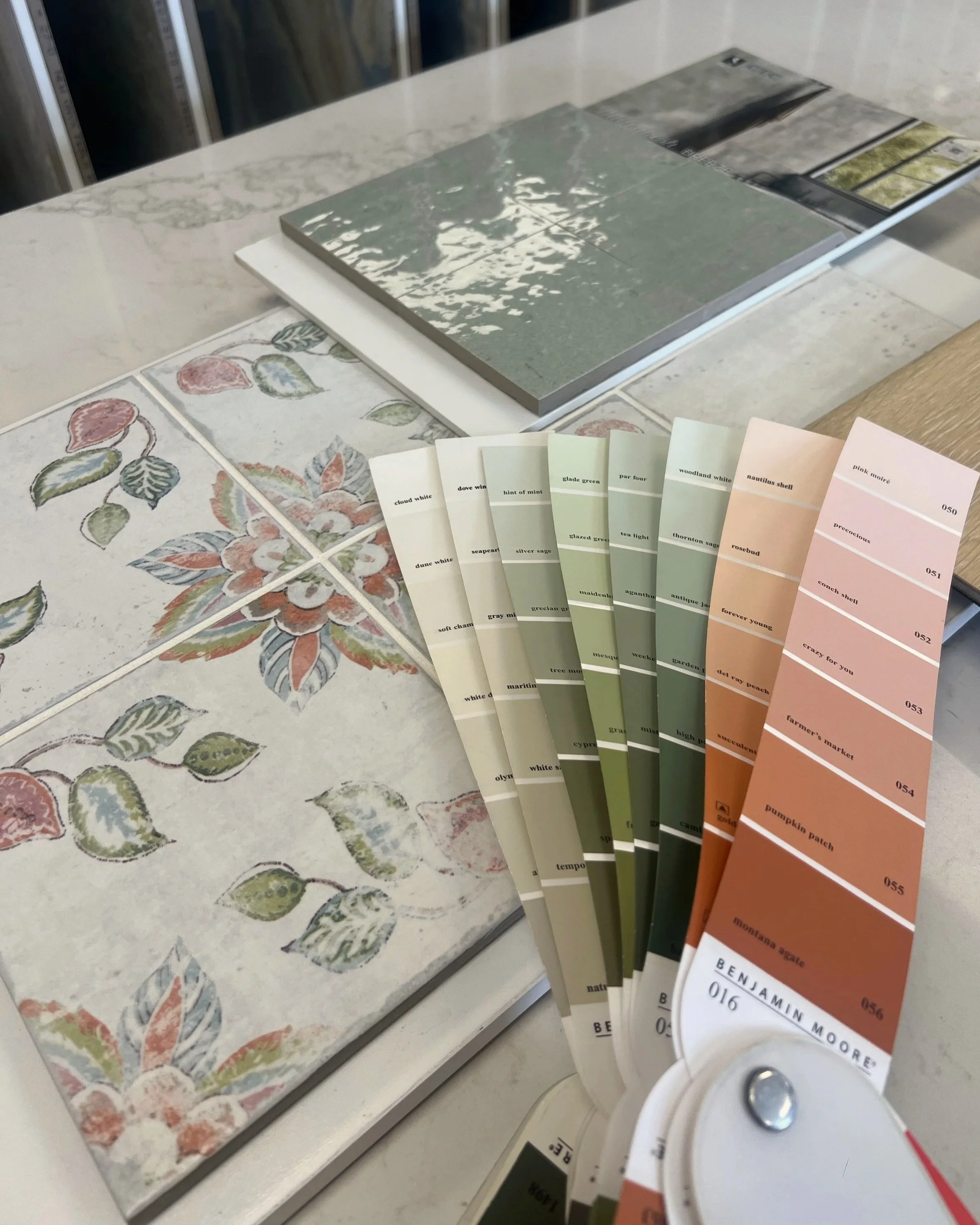 Paint color samples arranged in a fan shape with shades of green, orange, and pink, placed on a countertop with decorative tiles featuring floral and leaf patterns.