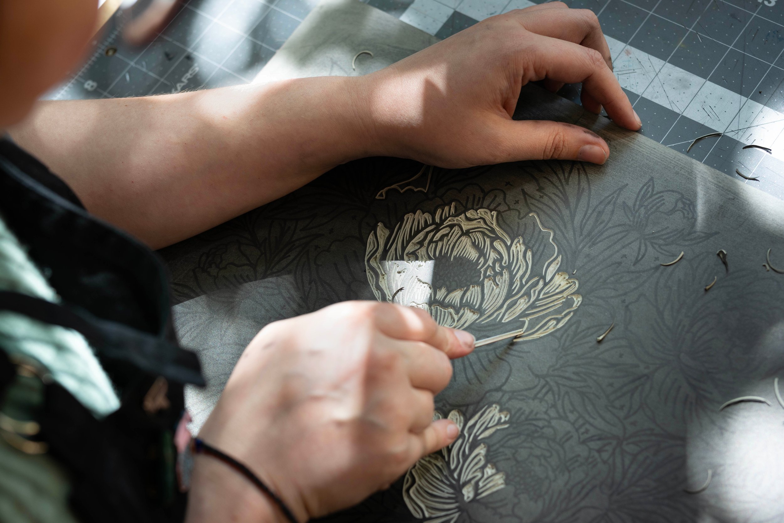 Close-up of a person's hand carving a detailed floral design into a dark linoleum surface.