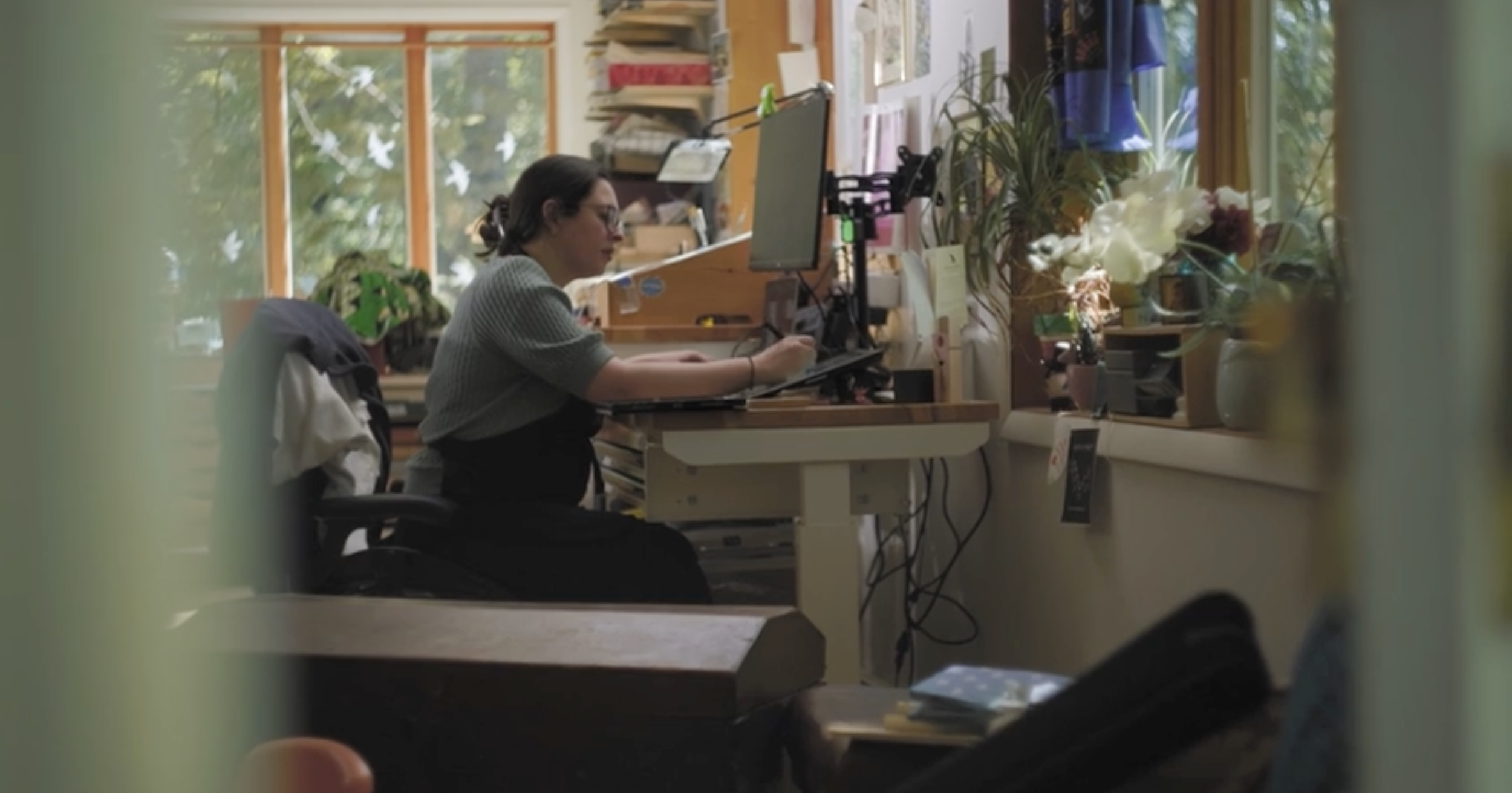 A woman working at a desk in a cozy, well-lit office with large windows, plants, and various office supplies.