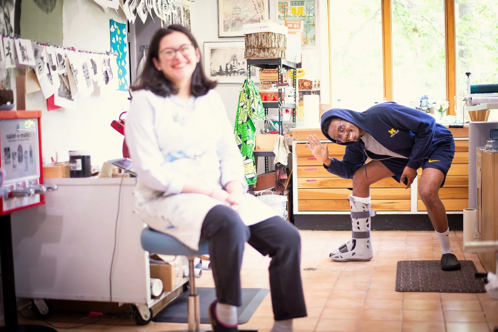 A woman sitting in a chair with a smile, wearing glasses and a white top, in a room decorated with various artwork and colorful items. A man, wearing a hoodie and shorts, with a big smile, is bending over with his foot in a protective boot, waving at the camera.